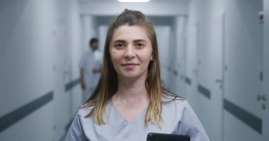 Kadın doktor modern klinik koridorunda duruyor. Profesyonel doktor dijital tableti tutuyor, gülümsüyor ve kameraya bakıyor. Hastane ya da tıp merkezi koridorundaki tıbbi personel ve hastalar.