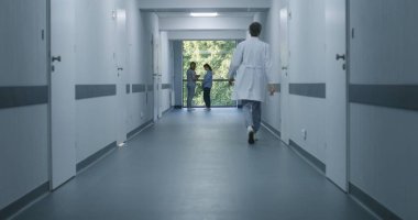 Klinik koridoru: Doktorlar ve profesyonel sağlık görevlileri yürüyor. Dijital tabletli hemşire pencerenin yanında duran yaşlı kadına geliyor. Modern hastane veya tıp merkezi koridorundaki tıbbi personel ve hastalar.