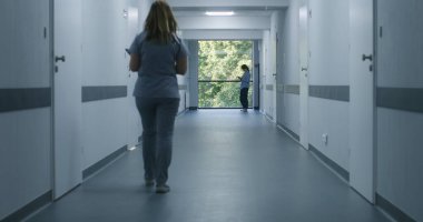 Klinik koridoru: Doktorlar ve profesyonel sağlık görevlileri yürüyor. Dijital tabletli hemşire pencerenin yanında duran kadın hastaya geliyor. Modern hastane veya tıp merkezi koridorundaki tıbbi personel ve hastalar.