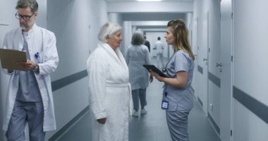 Kadın doktor, dijital tabletli hemşire ve hasta sohbeti. Doktor, prosedürlerden sonra yaşlı bir kadınla modern klinik koridorunda duruyor. Hastane ya da tıp merkezi koridorundaki hasta ve sağlık personeli.