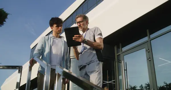 Mature doctor with digital tablet in hands stands with adult patient at clinic entrance and discuss his treatment. Health care specialist consults client or makes diagnosis. Physician works outside.