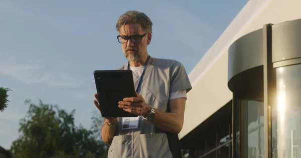 Mature doctor uses tablet computer, checks medical tests results. Medic in uniform stands outdoors at the modern hospital entrance. Clinic or medical center. Digital technologies in healthcare system.