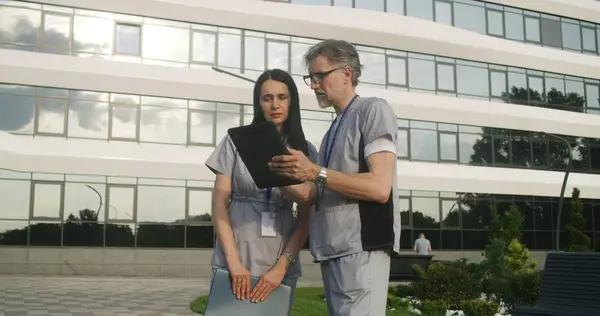 Two doctors stands outdoor and discuss patient treatment. Health care specialists look at client tests results using tablet. Woman helps colleague with making diagnosis. Medical staff work outside.