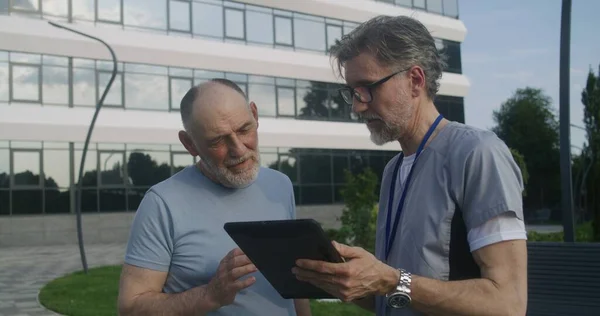 Mature doctor with digital tablet in hands stands with elderly patient outdoor and discuss his treatment. Health care specialist consults aged client or makes diagnosis. Medical staff work outside.