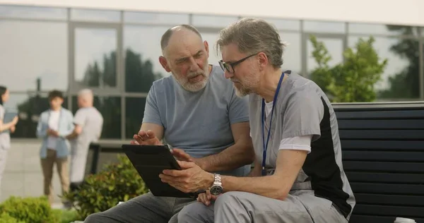 Mature doctor with tablet in hands sits with elderly patient on bench and consults him. Professional in medical uniform makes diagnosis to client using digital tablet. Medical staff work outdoor.