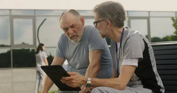 Mature doctor with tablet in hands sits with elderly patient on bench and consults him. Professional in medical uniform makes diagnosis to client using digital tablet. Medical staff work outdoor.