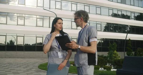 Two doctors stands outdoor and discuss patient treatment. Health care specialists look at client tests results using tablet. Woman helps colleague with making diagnosis. Medical staff work outside.