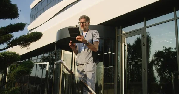 Mature doctor uses tablet computer, checks medical tests results. Medic in uniform stands outdoors at the modern hospital entrance. Clinic or medical center. Digital technologies in healthcare system.