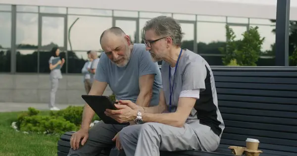 Mature doctor with tablet in hands sits with elderly patient on bench and consults him. Specialist in medical uniform discusses with man tests results using digital tablet. Medical staff work outdoor.