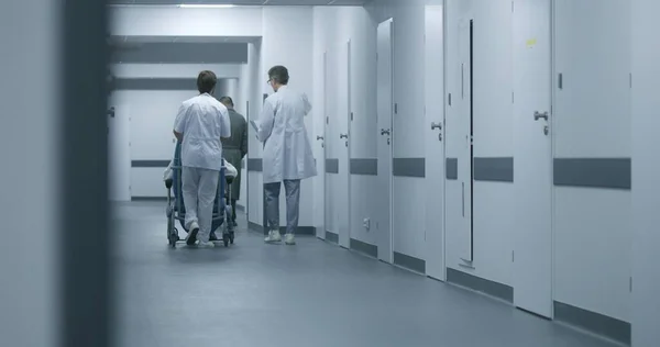 Back view of doctors pushing transfer wheelchair with patient walking along corridor. Medics talk, take person on procedures, therapy or to hospital room. Medical staff and patients in clinic hallway.