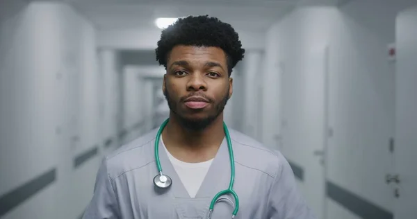 African American doctor stands in middle of medicine center hallway. Professional medic looks at camera and smiles. Caucasian doctor walks down the corridor with tests results. Medical staff at work.