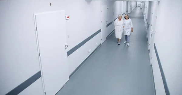 Bright clinic corridor: Female doctor and elderly woman walk down the hallway to hospital ward, talk about treatment. Medical staff work in modern medical facility. Security camera view. High angle.