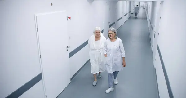 明るい診療所の廊下 女性医師と高齢女性が廊下を歩いて病院の病棟へ行き 治療について話します 医療スタッフは現代医療施設で働いています セキュリティカメラビュー 高角度 — ストック写真