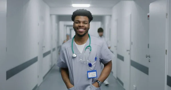 African American doctor looks at camera and smiles. Young doctor stands in middle of hospital corridor. Nurse transports senior patient in wheelchair in medical room. Medical staff at work in clinic.