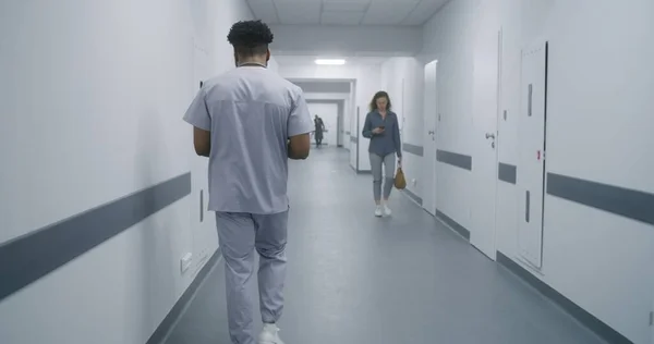 African American health care specialist walks hospital hallway. Male doctor goes on appointment with patients. Multi ethnic medical personnel at work in modern clinic.