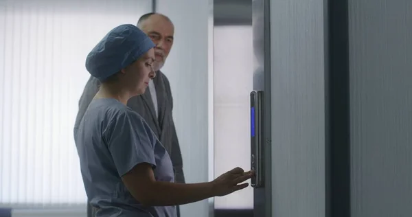 Nurse stands near lift door, talks with patient and comes in elevator with him. Healthcare specialist at work in modern medical center. Medical worker and aged patient in clinic hallway. Close up.