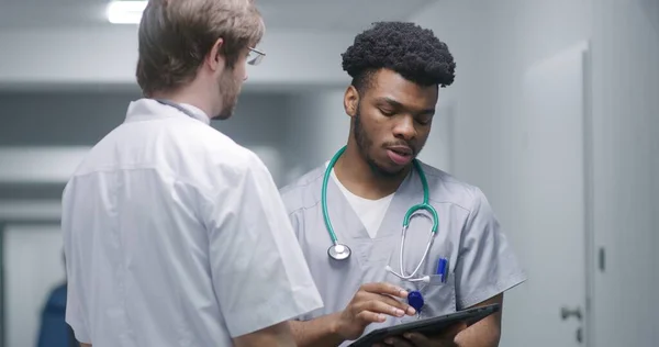 African American doctor stands in medical center hallway and works on his digital tablet. Healthcare specialist debates with colleague about test results in clinic corridor. Medical staff at work.