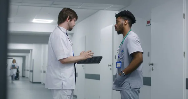 African American doctor stands in the middle of medic center hallway with his colleague. Young doctor discusses with other medic patient treatment and shows tests results on digital tablet computer.