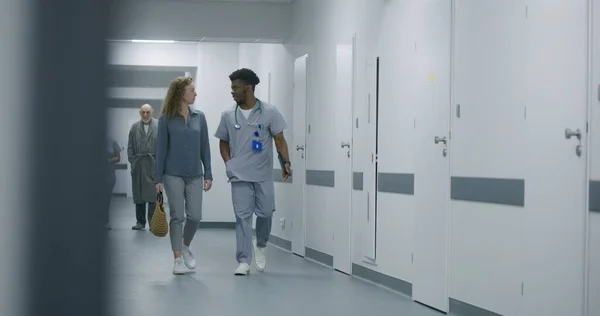 African American doctor walks down the medical center hallway with digital tablet computer and his patient. Healthcare specialist talks with woman about medical test results in hospital corridor.