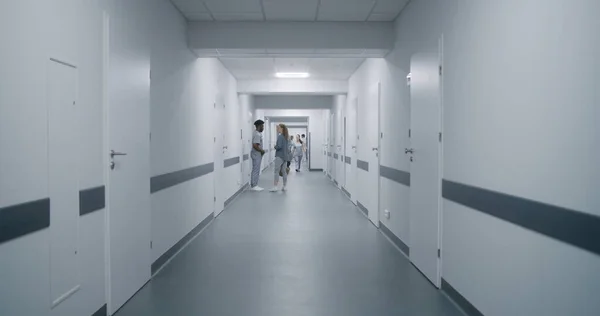 Woman goes medical center corridor, talks to African American doctor. Nurse and elderly patient walk clinic hallway, discuss tests results. Multiracial medical staff in clinic.