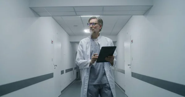 Female medic goes to medical room with old patient. Elderly doctor opens door with key card and comes in with her patient. Healthcare specialist walks down the corridor and work on digital tablet.