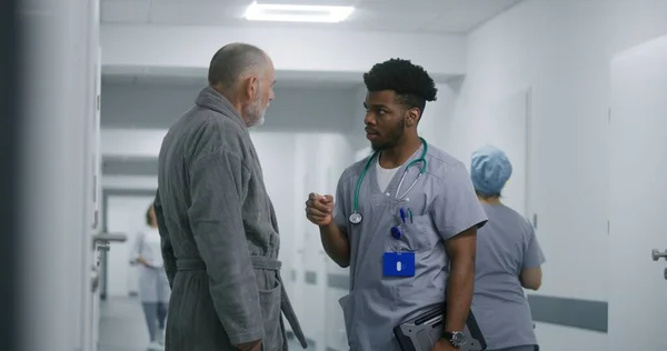 African American doctor asks elderly patient about his health in medical center hallway. Senior man stands in clinic corridor and talks with professional medic about treatment. Medical staff at work.
