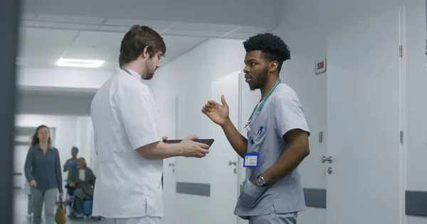 Professional doctor stands in hospital corridor, uses digital tablet, shows MRI or CT brain scan results to his African American colleague. Medics debates about treatment of patient in clinic hallway.