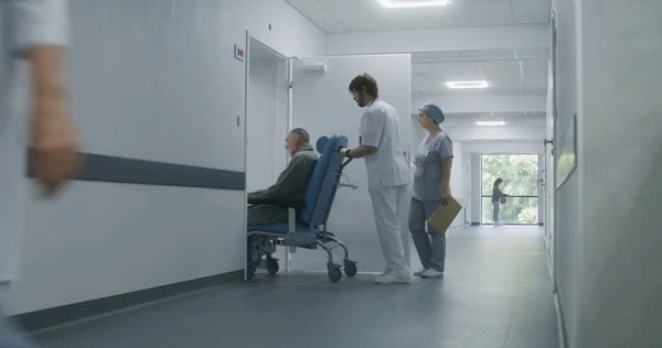 Nurse opens door to medical room with key card in hospital hallway. Adult doctor transports elderly patient on wheelchair in cabinet for medical procedures. Medical staff at work in modern clinic.