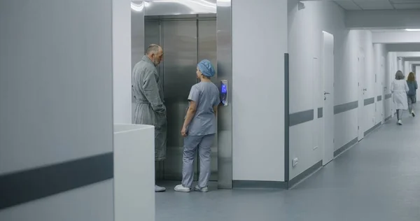 Nurse in uniform walks hospital corridor with aged man and comes to lift door. Female healthcare specialist meets elderly patient in medical center hallway. Medical personnel work in modern clinic.