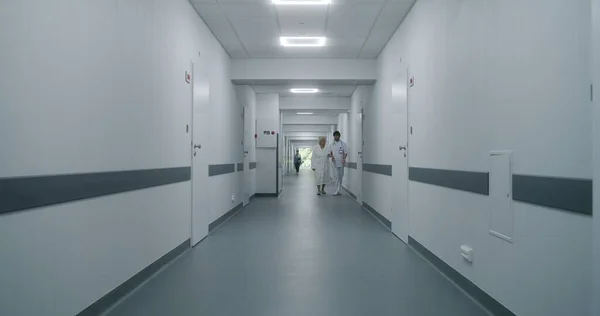 Mature doctor walks down hospital corridor with digital tablet computer to medical room. Professional medic stops to talk to young colleague and old patient. Medical staff at work in medical center.