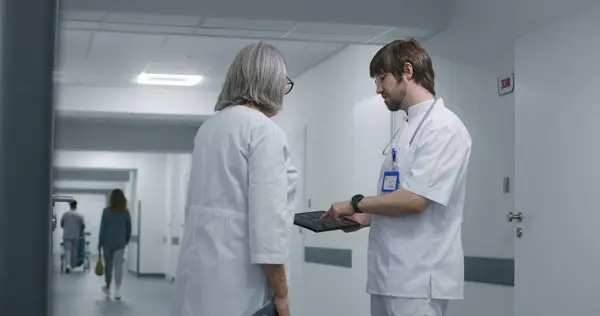 Young doctor consults about MRI or CT scanning tests with mature female medic in medical center hallway. Professional doctor uses digital tablet computer. Medical staff at work in modern clinic.