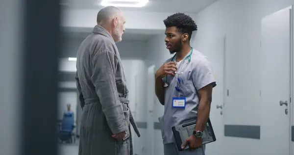 African American doctor asks elderly patient about his health in middle of hospital hallway. Young medic discusses medical tests results with old man. Nurse transports wheelchair to medical cabinet.