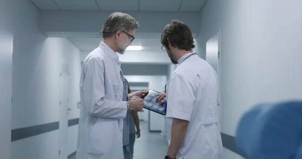 成熟した医師は デジタルタブレットを使用して同僚にMri Ct脳スキャン画像を示しています 専門医が立ってクリニックの廊下を歩き 仕事について話し合います 医療スタッフ 病院の廊下の患者さん — ストック写真