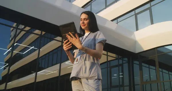 Female doctor consults patient during online appointment using digital tablet. Medic in uniform stands outdoors near hospital. Medical staff of clinic or medical center. Modern healthcare system.