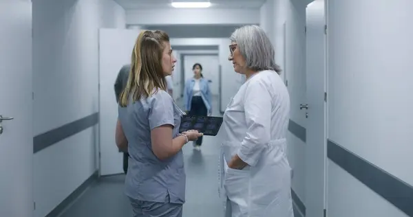 看護師は デジタルタブレットを使用して女性医師にMriまたはCt脳スキャン画像を示しています 医学は現代の診療所の廊下に立って歩き 仕事について話し合います 病院または医療センターの廊下の医療スタッフ — ストック写真