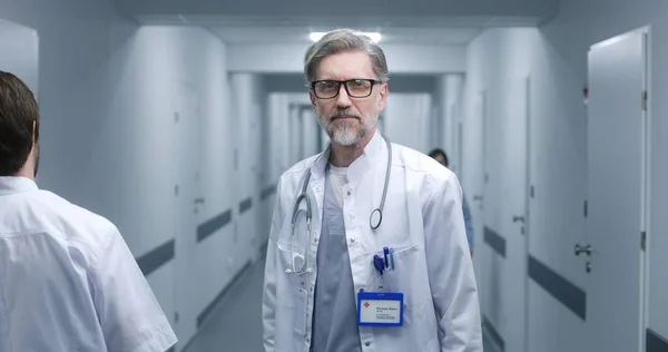 Mature doctor stands in modern clinic corridor. Professional medic puts on glasses, smiles, looks at camera. Medical staff and patients in background in hospital or medical center hallway. Portrait.
