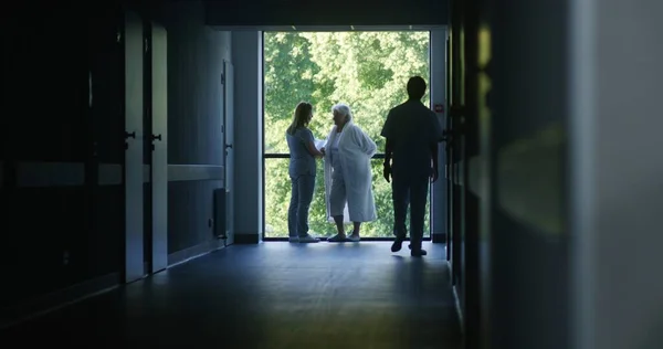 Clinic corridor: Doctors and professional medics walk. Nurse with papers comes to elderly female patient standing near window. Medical staff and patients in dark hospital or medical center hallway.