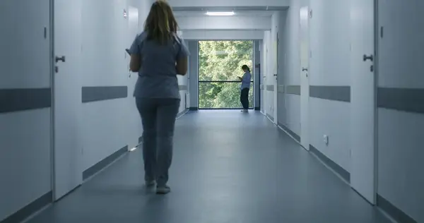 Clinic corridor: Doctors and professional medics walk. Nurse with digital tablet comes to female patient standing near window. Medical staff and patients in modern hospital or medical center hallway.
