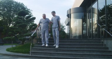 Modern hastanenin yakınında üniformalı profesyonel doktorlar yürüyor. Olgun sıhhiyeci dijital tablet tutuyor, iş arkadaşlarıyla konuşuyor. Klinik ya da Tıp Merkezi 'nin tıbbi personeli. Sağlık hizmetleri sistemi. Yavaş çekim.