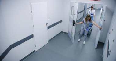 Klinik koridoru: Doktorlar, profesyonel sağlık görevlileri ve hastalar yürüyor. Erkek hemşire tekerlekli sandalye sürüyor. Yaşlı adam hastane odasına gider. Modern tıbbi tesiste tıbbi personel çalışıyor. Yüksek açı.