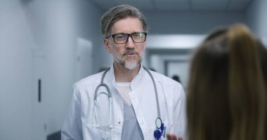 Olgun doktor modern klinik koridorunda duruyor, bayan meslektaşı ya da hastasıyla konuşuyor. Sağlık uzmanı tedaviyi ya da tanı sonuçlarını tartışır. Hastane koridorunda tıbbi personel ve hasta var..