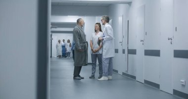 Olgun doktor, hemşire ve hastayla klinik koridorunda duruyor. Profesyonel doktor kadın meslektaşı ve yaşlı kadınla konuşuyor, dijital tablet kullanıyor. Hastane koridorunda tıbbi personel ve hasta var..