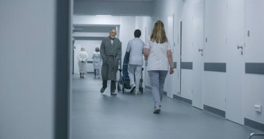 Olgun doktor, hemşire ve hastayla klinik koridorunda duruyor. Profesyonel doktor kadın meslektaşı ve yaşlı kadınla konuşuyor, dijital tablet kullanıyor. Hastane koridorunda tıbbi personel ve hasta var..