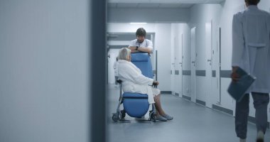 Erkek doktor, klinik koridorunda tekerlekli sandalyedeki kadın hastayla birlikte duruyor. Ameliyathane yakınındaki ameliyattan önce doktor yaşlı bir kadınla konuşur. Sağlık personeli, hastanedeki ya da sağlık merkezindeki hastalar.