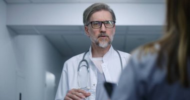 Olgun doktor modern klinik koridorunda duruyor, bayan meslektaşı ya da hastasıyla konuşuyor. Sağlık uzmanı tedaviyi ya da tanı sonuçlarını tartışır. Hastane koridorunda tıbbi personel ve hasta var..