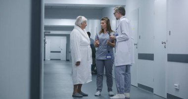 Hemşire ve yaşlı kadın prosedürlerden sonra klinik koridorunda yürüyorlar. Erkek doktor iş arkadaşı ve hastasına gelir, test sonuçlarını dijital tablet kullanarak gösterir. Hastane koridorunda tıbbi personel ve hasta var..