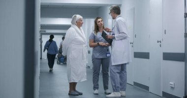 Hemşire ve yaşlı kadın prosedürlerden sonra klinik koridorunda yürüyorlar. Erkek doktor iş arkadaşı ve hastasına gelir, test sonuçlarını dijital tablet kullanarak gösterir. Hastane koridorunda tıbbi personel ve hasta var..