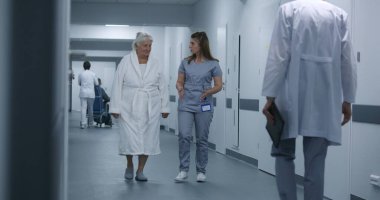 Hemşire ve yaşlı kadın prosedürlerden sonra klinik koridorunda yürüyorlar. Erkek doktor iş arkadaşı ve hastasına gelir, test sonuçlarını dijital tablet kullanarak gösterir. Hastane koridorunda tıbbi personel ve hasta var..