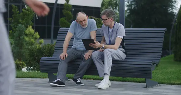 Mature doctor with tablet in hands sits with elderly patient on the bench and consults him. Medic in uniform prescribes medicines to client using digital tablet computer. Medical staff work outdoor.