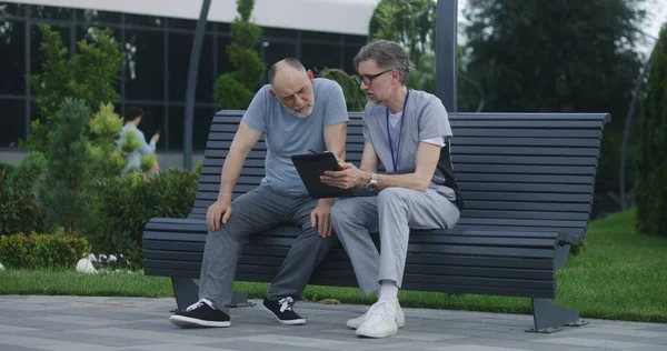 Mature doctor with tablet in hands sits with elderly patient on the bench and consults him. Medic in uniform prescribes medicines to client using digital tablet computer. Medical staff work outdoor.